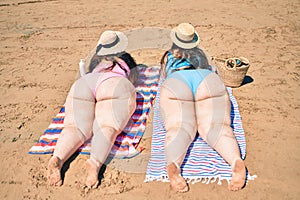 Backview of two plus size overweight sisters twins women relaxing lying on a towel at the beach on summer holidays