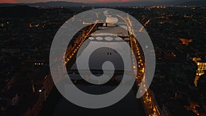 Backwards fly above Arno river at dusk. Waterfront streets and bridges lit by streetlights. Tilt up reveal colorful sky