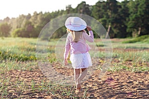 Backwards view of little girl in dress and white straw hat walking barefoot on sand at the sunset