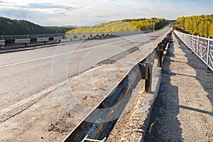 Bad asphalt road going over the bridge, fences along the road, forest and mountains