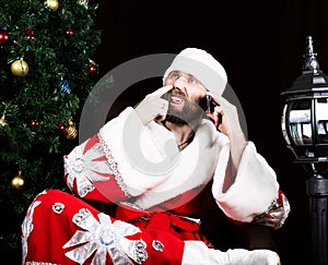 Bad brutal Santa Claus dissatisfied talking phone and picking his nose, on the background of Christmas tree