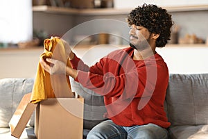 Bad delivery service concept. Dissatisfied indian man unpacking cardboard box with clothes