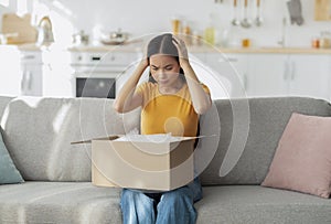 Bad delivery service. Dissatisfied asian lady sitting on sofa with cardboard box, received parcel with wrong item