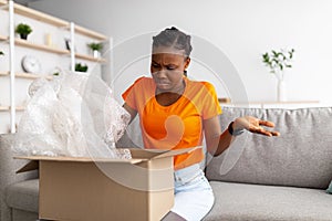 Bad delivery service, online shopping. Irritated black woman unboxing cardboard parcel, receiving damaged item at home