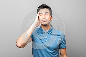 Only bad news. exhausted adult man in blue shirt with headache holding his hand over his temple standing against grey