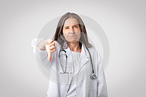 Bad service. Senior woman doctor in white coat with stethoscope showing thumb down sign on gray background