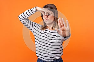 Bad smell. Portrait of young woman with brown hair in long sleeve striped shirt. indoor studio shot isolated on orange background