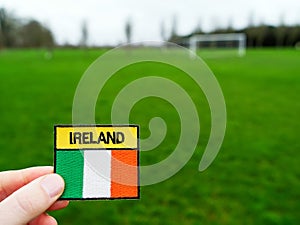 Badge with Irish National flag and inscription Ireland in focus, green grass football field and goal posts out of focus. Soccer