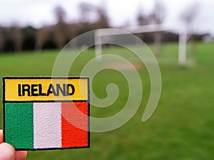 Badge with Irish National flag and inscription Ireland in focus, green grass football field and goal posts out of focus. Soccer