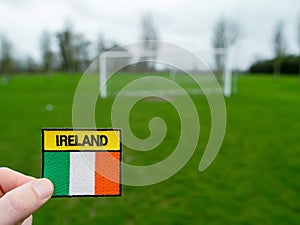 Badge with Irish National flag and inscription Ireland in focus, green grass football field and goal posts out of focus. Soccer