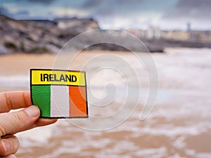 Badge with sign Ireland and Irish flag in focus. Lahinch ocean town out of focus in the background. Travel concept