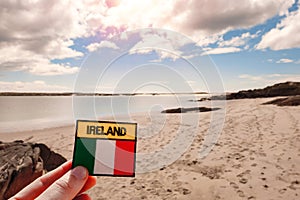 Badge with sign Ireland and National flag in focus. Gurteen beach out of focus, county Galway, Ireland. Warm sunny day. Cloudy sky