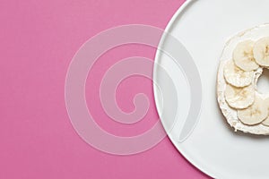 Bagel with cream cheese and banana on a white plate on pink
