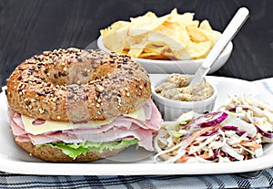 Bagel ham, cheese and lettuce sandwich with a side a spicy mustard and coleslaw.