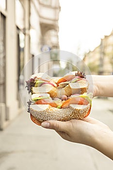 Bagel with prosciutto, cream cheese in woman hand. Holding a Sandwich with lettuce for breakfast on street