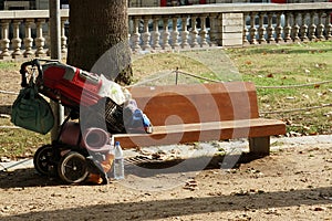 Baggage trolley with sleeping bag and hand luggage of a homeless person leaned against wooden bench.