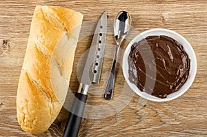 Baguette, knife, spoon, bowl with chocolate cheese on wooden table. Top view