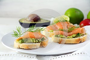 Baguette sandwiches with smoked salmon and avocado cream or guacamole as healthy party canapes on a white plate, ingredients in t