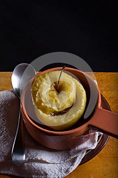 A baked apple in a clay pot on a linen cloth and wooden desk against black background