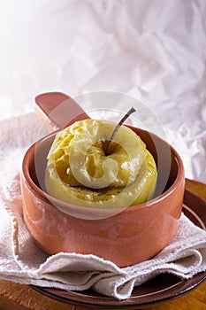 A baked apple in a clay pot on a linen cloth and wooden desk against white background