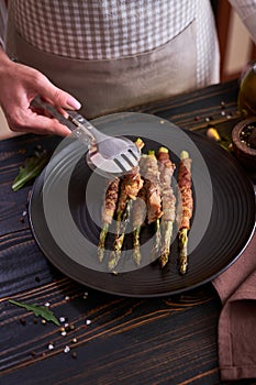 baked aspargus covered with bacon on a black ceramic plate on wooden kitchen table