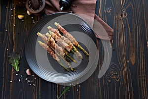 baked aspargus covered with bacon on a black ceramic plate on wooden kitchen table