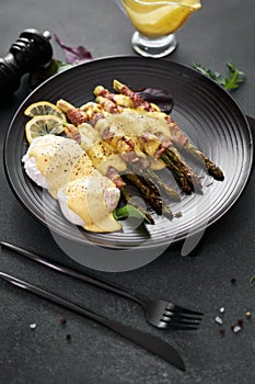 baked aspargus covered with bacon and poached eggs on a black ceramic plate