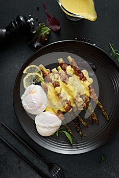 baked aspargus covered with bacon and poached eggs on a black ceramic plate