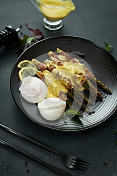 baked aspargus covered with bacon and poached eggs on a black ceramic plate