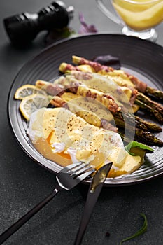baked aspargus covered with bacon and poached eggs on a black ceramic plate