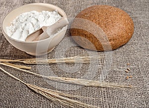 Baked bread, a bowl of flour and a sheaf of wheat against a background of burlap