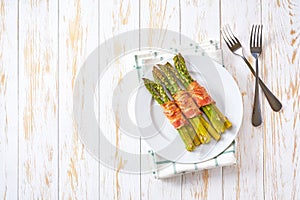 Baked bundles of green asparagus wrapped in bacon in a ceramic plate on a white wooden table, top view