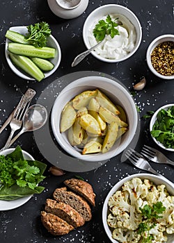 Baked cauliflower and potatoes, fresh cucumbers and herbs, sauces and spices on a dark stone background. Healthy vegetarian lunch