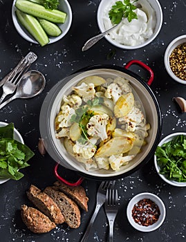 Baked cauliflower and potatoes in one pot, fresh cucumbers and herbs, sauces and spices on a dark stone background. Healthy