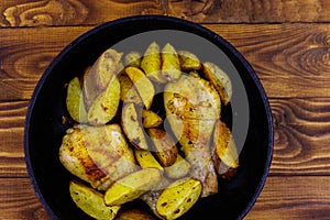 Baked chicken drumsticks with potatoes in in cast-iron frying pan on wooden table. Top view