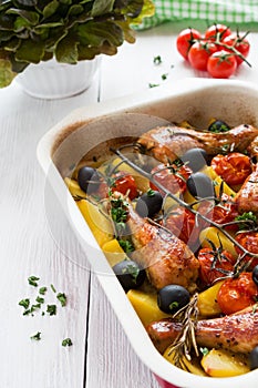 Baked chicken drumsticks in red dish. Cooked with cherry tomatoes, black olives, rosemary and potatoes. White wooden table