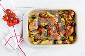 Baked chicken drumsticks in red dish. Cooked with cherry tomatoes, black olives, rosemary and potatoes. White wooden table