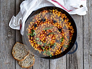 Baked chickpea with vegetables, olives and sun-dried tomatoes in iron pan, frying pan on wooden table. Vegan vegetarian