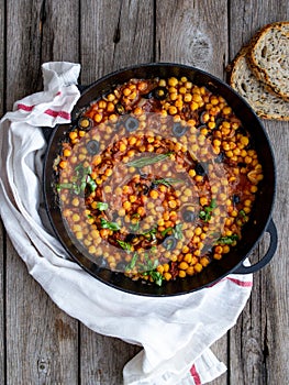 Baked chickpea with vegetables, olives and sun-dried tomatoes in iron pan, frying pan on wooden table. Vegan vegetarian