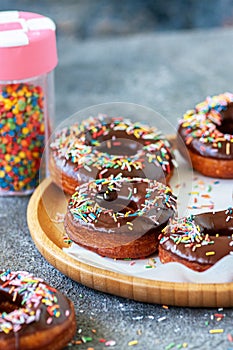 Baked chocolate doughnuts with colorful sprinkles on a wooden plate. Gray background