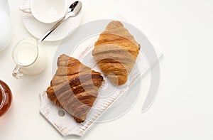 Baked croissants, white ceramic teapot and empty cup and saucer, jar of honey on a white table, top view. Breakfast