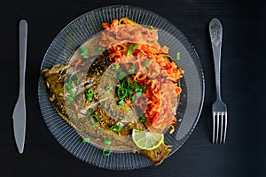 Baked fish flounder with lemon, carrot and spicy herbs, closeup on black background. Delicious fish dish with vegetables for