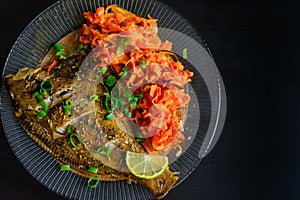 Baked fish flounder with lemon, carrot and spicy herbs, in a plate closeup on a black background. Delicious fish dish with