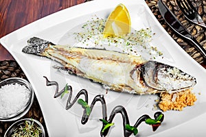 Baked fish with lemon and greens on plate on wooden background.