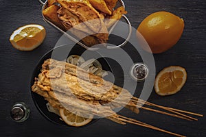 Baked fish on skewers in a black plate, french fries and lemon on a wooden table.