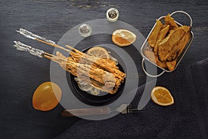 Baked fish on skewers in a black plate, french fries and lemon on a wooden table.