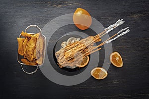 Baked fish on skewers in a black plate, french fries and lemon on a wooden table.