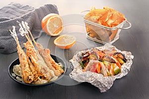 Baked fish on skewers in a black plate, vegetables in foil, french fries and lemon on a wooden table