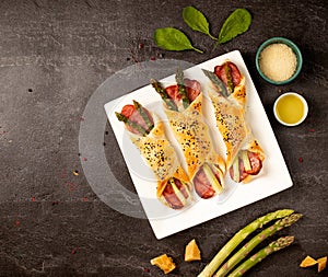 Baked green asparagus with ham and cheese in puff pastry sprinkled with sesame seeds and green leaves. Light gray stone