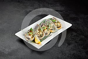 Baked mussels with cheese on plate on dark concrete background. Appetizer of mussels and lemon in minimal style on black table.